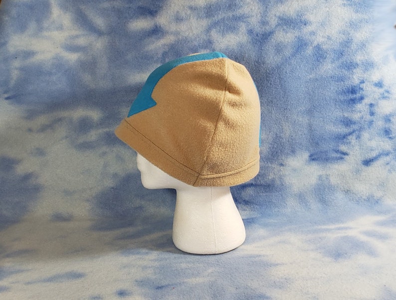 May include: A tan fleece beanie with a blue arrow design on the front. The beanie is on a white mannequin head.