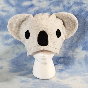 May include: A gray plush koala hat with black felt ears and eyes. The hat is on a white mannequin head.