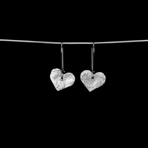 May include: A pair of silver-toned heart-shaped earrings suspended from a thin silver chain. Each heart features a detailed, textured surface with intricate swirling patterns. The earrings hang vertically from simple, slender metal hooks.