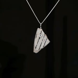 May include: A silver-toned snake chain necklace featuring a pendant made from a triangular, textured ceramic shard. The off-white shard displays thin, vertical black stripes and is secured to the chain with a coiled silver wire bail.