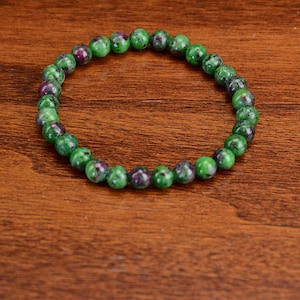 May include: A circular beaded bracelet composed of polished, spherical stones. The beads feature a mottled pattern of vibrant green and deep purple-red hues, strung together on an elastic cord to form a continuous, flexible loop for the wrist.