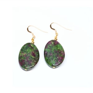 May include: A pair of drop earrings featuring oval-shaped, polished stones with a mottled pattern of deep forest green and vibrant magenta. Each stone is suspended from a thin, gold-toned metal hook wire, connected by a small gold-toned bead.