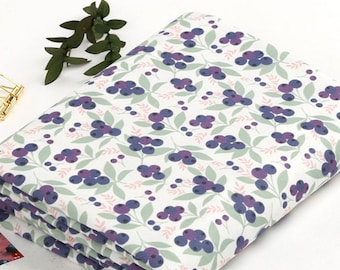 May include: Folded fabric with a white background and a repeating pattern of purple blueberries, green leaves, and pink accents. The fabric is stacked, showing the pattern on the top layer. A sprig of green leaves is placed on the top left.