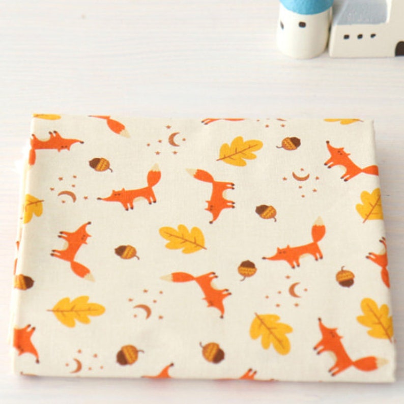May include: White fabric with a repeating pattern of orange foxes, yellow leaves, and brown acorns. The fabric has a subtle texture.