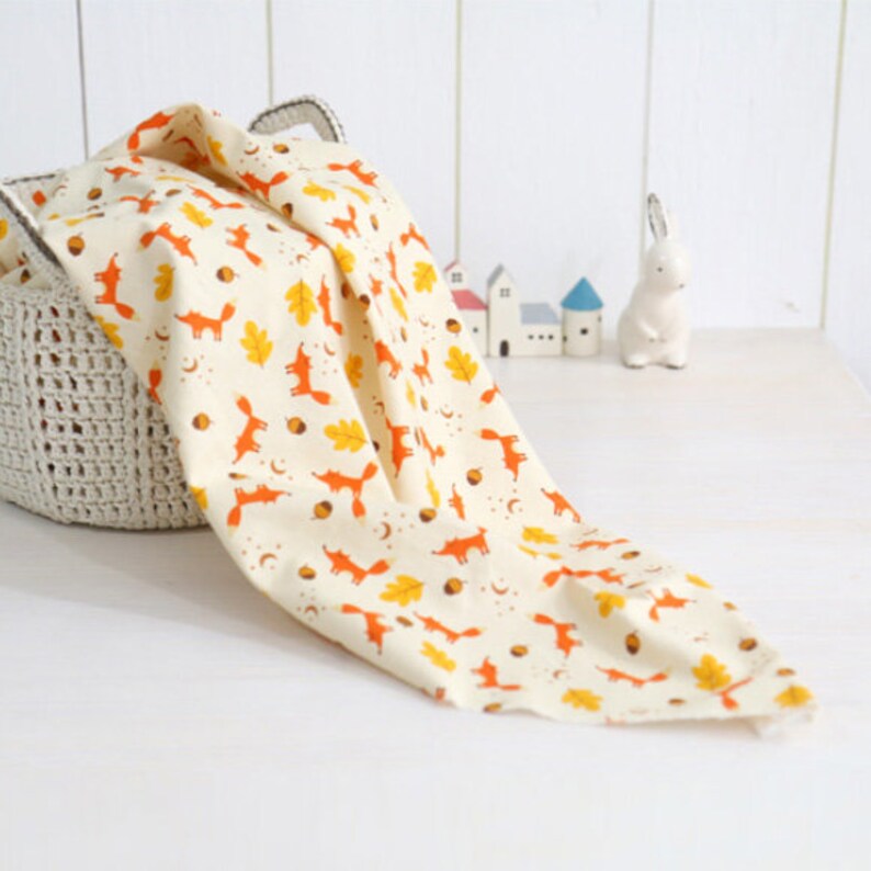 May include: A cream-colored fabric with a repeating pattern of orange foxes, yellow leaves, and brown acorns. The fabric is draped over a crocheted basket.