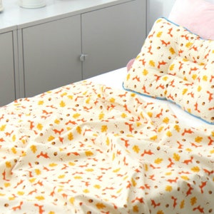 May include: A white cotton blanket with a repeating pattern of orange foxes and yellow leaves. The blanket is on a bed with a white pillow with the same pattern.