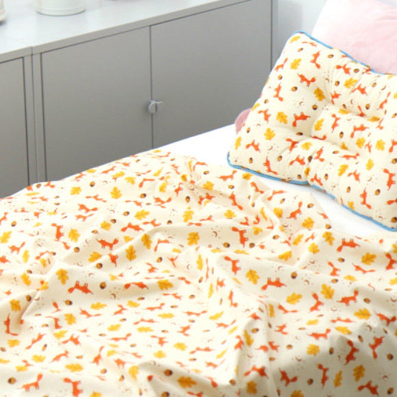 May include: A white cotton blanket with a repeating pattern of orange foxes and yellow leaves. The blanket is on a bed with a white pillow with the same pattern.