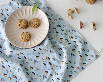 May include: A light blue cotton tea towel with a peanut pattern. The towel is folded and placed on a white surface with a plate of cookies and peanuts.