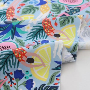 May include: Light blue fabric with a vibrant print featuring swimmers, lemons, and tropical foliage. The design includes elements of red, yellow, green, and blue, creating a summery and playful aesthetic. The fabric is folded, showcasing the pattern's repeat.