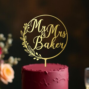 May include: A two-tiered burgundy cake with a gold cake topper that reads "Mr & Mrs Baker" in a circular design. The cake sits on a gold cake stand, surrounded by flowers and champagne glasses, perfect for a wedding.