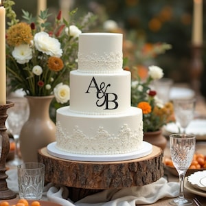 May include: A three-tiered white wedding cake with lace detailing and the initials "A & B" on the middle tier. The cake is on a wooden cake stand and surrounded by candles, flowers, and other wedding decorations.