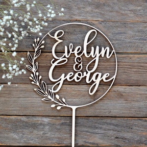 May include: A wooden cake topper with the names "Evelyn & George" in elegant script. The names are inside a circle, with a leafy design on the left side. The topper has a pointed stake for insertion into a cake.