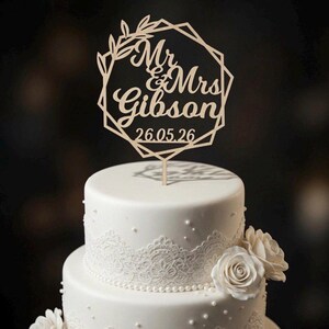 Op de afbeelding: Een lichtgekleurde houten taarttopper met een geometrisch frame met bladaccenten, de tekst Mr & Mrs Gibson en de datum 26.05.26. De topper staat op een witte taart met twee lagen, versierd met kanten randen, parelkralen en witte rozen.