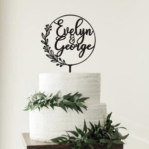 May include: A black cake topper with the names "Evelyn & George" in a circular design, surrounded by a floral wreath. The topper is placed on a two-tiered white cake decorated with greenery.
