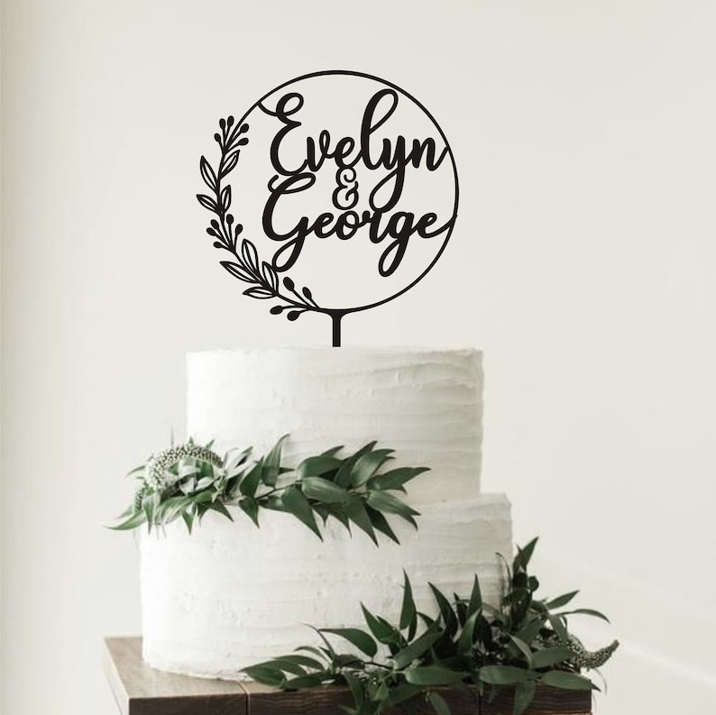May include: A black cake topper with the names "Evelyn & George" in a circular design, surrounded by a floral wreath. The topper is placed on a two-tiered white cake decorated with greenery.
