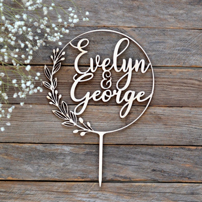 May include: A wooden cake topper with the names "Evelyn & George" in elegant script. The names are inside a circle, with a leafy design on the left side. The topper has a pointed stake for insertion into a cake.
