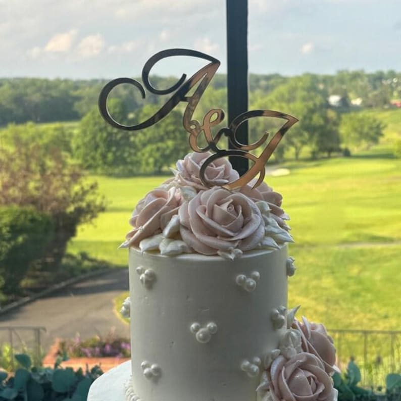 May include: A wedding cake with light pink roses and a gold-colored topper with the letters "A & J". The cake is white with decorative pearl-like accents. The background shows a green landscape with trees and a golf course.