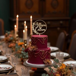 May include: A two-tiered burgundy cake with gold accents and floral decorations. A wooden cake topper with the names "Allison & Calum" is placed on top. The cake sits on a decorative cake stand, surrounded by candles and table settings.