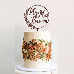May include: A wedding cake with buttercream frosting and floral decorations. A brown wooden cake topper reads "Mr & Mrs Brown" inside a circle with leaf accents. The cake sits on a white cake board.