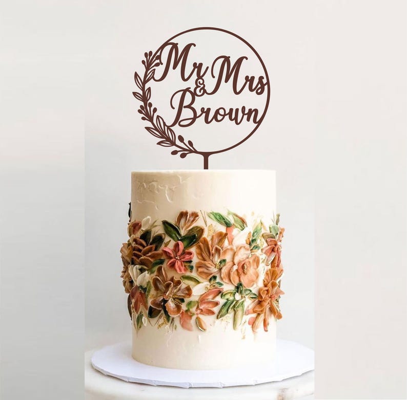 May include: A wedding cake with buttercream frosting and floral decorations. A brown wooden cake topper reads "Mr & Mrs Brown" inside a circle with leaf accents. The cake sits on a white cake board.