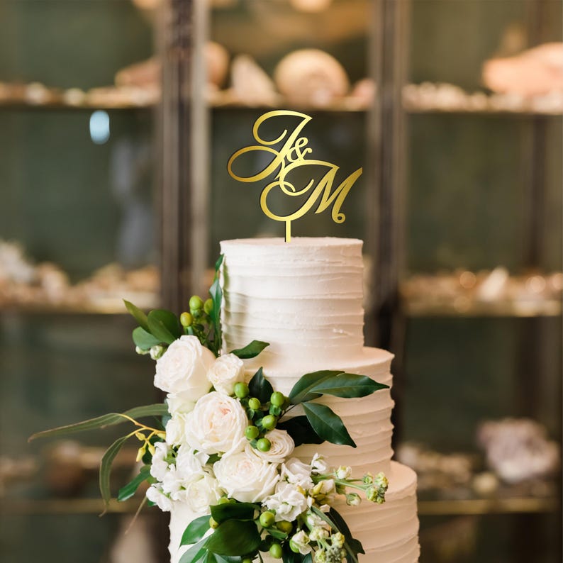 May include: A three-tiered white wedding cake with a gold-colored topper featuring the letters "J&M". The cake is decorated with white roses, greenery, and small green berries. The background is a blurred interior.