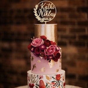 May include: A three-tiered cake featuring a bottom layer with a botanical floral print, a middle layer in light pink with gold leaf accents and fresh red and pink roses, and a top gold layer topped with a circular gold cake topper reading Kathi & Niklas 08.08.26.