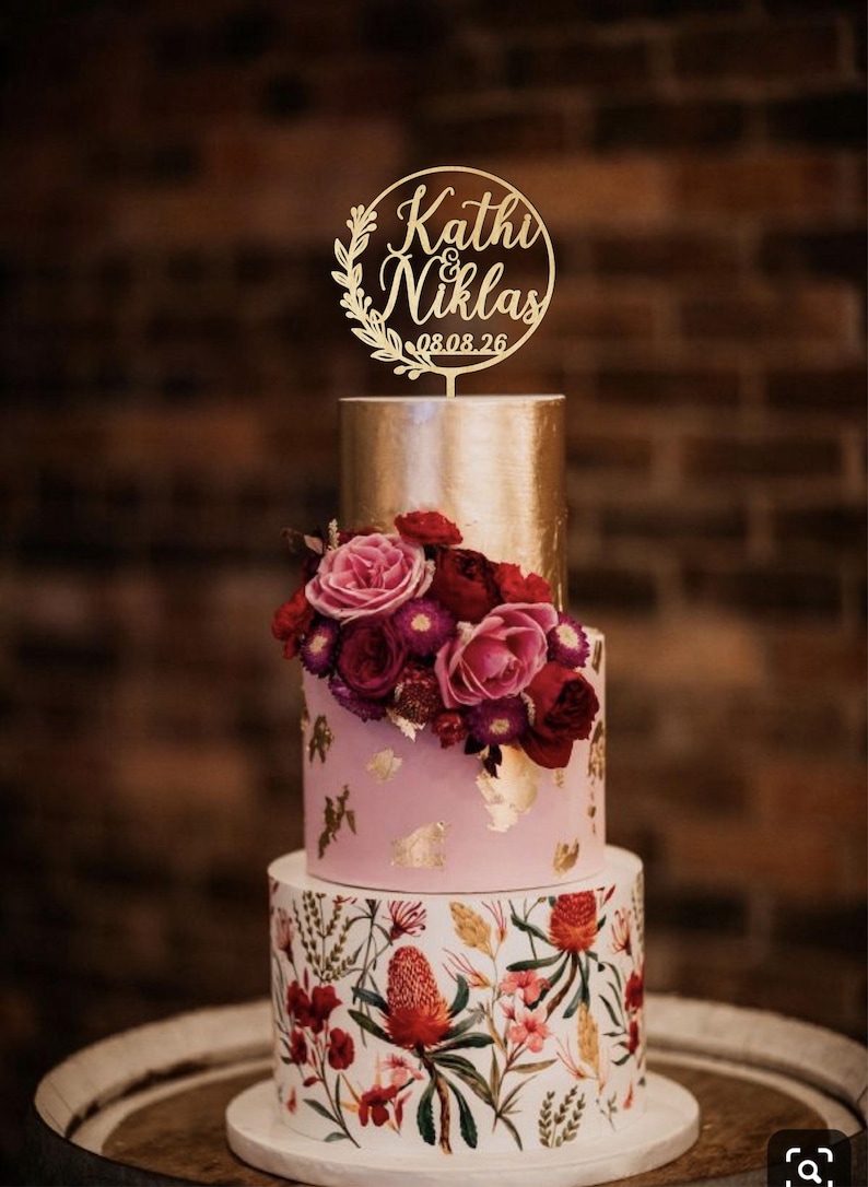 May include: A three-tiered cake featuring a bottom layer with a botanical floral print, a middle layer in light pink with gold leaf accents and fresh red and pink roses, and a top gold layer topped with a circular gold cake topper reading Kathi & Niklas 08.08.26.