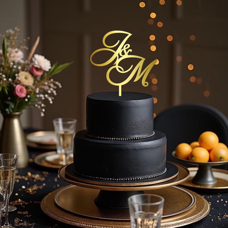 May include: A two-tiered black cake with a gold topper featuring the letters "J & M" in an elegant script. The cake sits on a gold cake stand, with a dark background and bokeh lights.