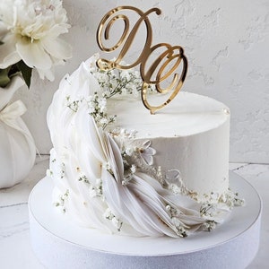 May include: A white frosted cake decorated with white fabric and small white flowers. A gold-colored "D&D" cake topper sits atop the cake. The cake is on a white cake stand, with white flowers in the background.