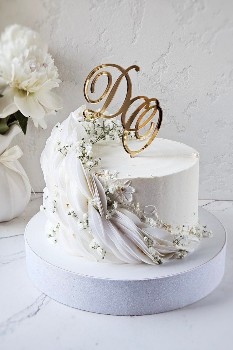 May include: A white frosted cake decorated with white fabric and small white flowers. A gold-colored "D&D" cake topper sits atop the cake. The cake is on a white cake stand, with white flowers in the background.