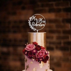 May include: A two-tiered wedding cake with a gold top tier and a pink bottom tier decorated with gold leaf and flowers. A white cake topper reads "Mr & Mrs Robertson" in a circular design with floral accents.