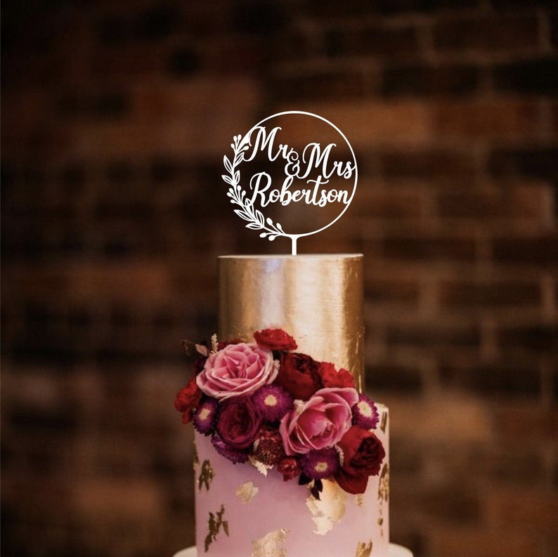 May include: A two-tiered wedding cake with a gold top tier and a pink bottom tier decorated with gold leaf and flowers. A white cake topper reads "Mr & Mrs Robertson" in a circular design with floral accents.