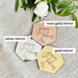May include: Three hexagonal mirrors in rose gold, silver, and gold. Each mirror has the text "MR&MRS Heikens" and a floral design. The mirrors are displayed on a neutral linen surface with floral accents.