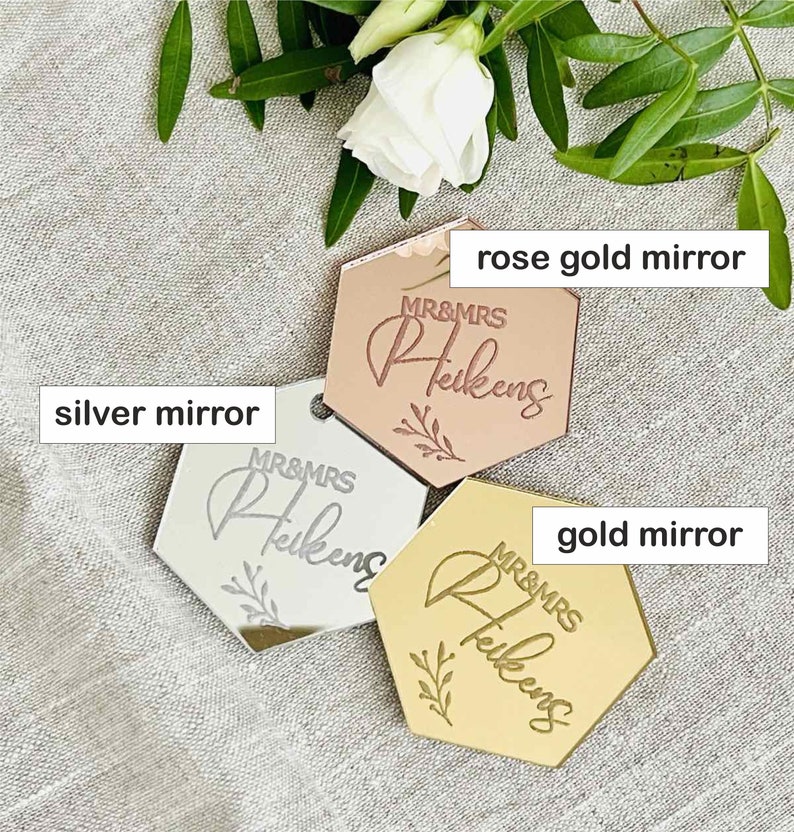 May include: Three hexagonal mirrors in rose gold, silver, and gold. Each mirror has the text "MR&MRS Heikens" and a floral design. The mirrors are displayed on a neutral linen surface with floral accents.