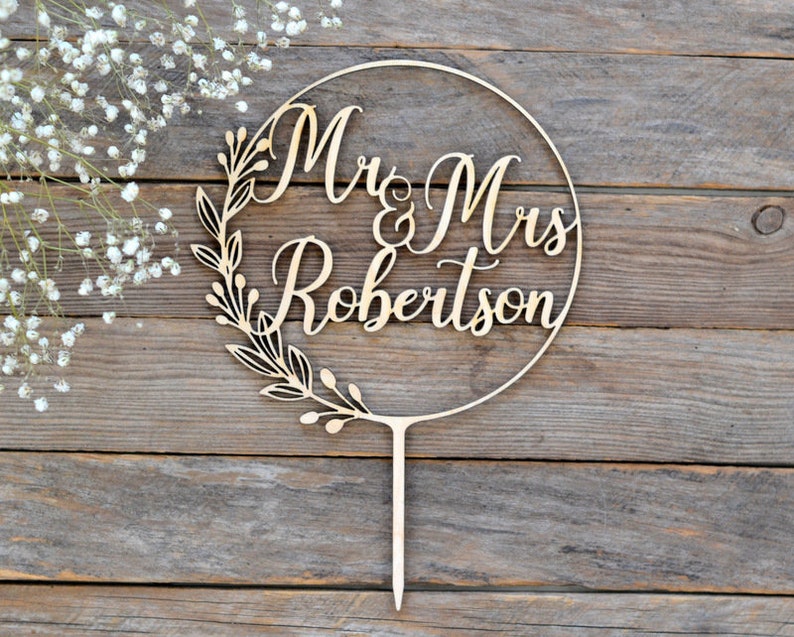 May include: A wooden cake topper with the words "Mr & Mrs Robertson" in elegant script, encircled by a floral wreath. The topper is light brown and has a stake for insertion into a cake. The background is a rustic wooden surface.
