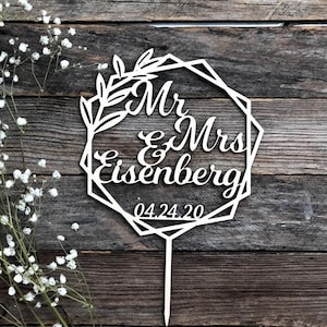 May include: White wooden cake topper with a geometric frame and floral design. The topper says "Mr & Mrs Eisenberg" and "04.24.20".