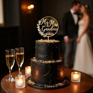 May include: A two-tiered black wedding cake with a wooden topper that reads "Mr & Mrs Gardere 10.14.23". The cake is decorated with a textured frosting and crumbs. Two champagne flutes and lit candles are on the table.