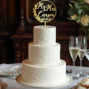 May include: A three-tiered white wedding cake with lace detailing and a gold cake topper. The topper reads "Mr & Mrs Carson 03.29.25" in a circular design with floral accents. The cake sits on a white pedestal cake stand.