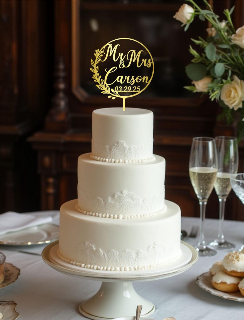 May include: A three-tiered white wedding cake with lace detailing and a gold cake topper. The topper reads "Mr & Mrs Carson 03.29.25" in a circular design with floral accents. The cake sits on a white pedestal cake stand.