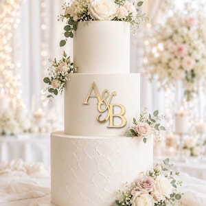 May include: A three-tier white wedding cake featuring a textured bottom layer and a smooth middle layer adorned with a gold-coloured monogram topper displaying the letters A & B. The cake is decorated with clusters of white and pale pink roses and greenery.