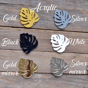 May include: A collection of acrylic monstera leaf cutouts in various colors, including gold, silver, black, and white. The words "Acrylic", "Gold", "Silver", "Black", "White", "Gold mirror", and "Silver mirror" are written in cursive.
