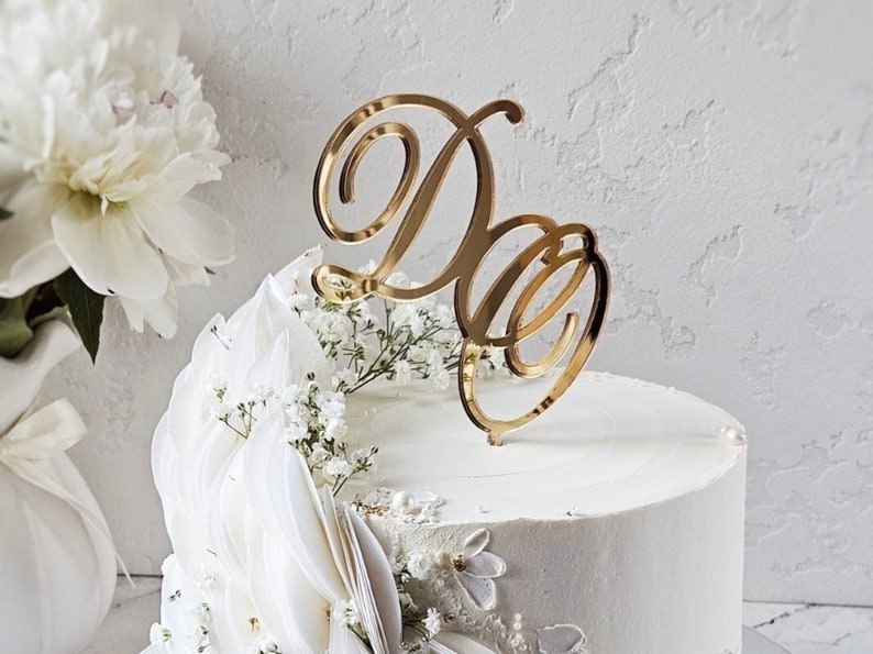 May include: A gold-colored cake topper with the intertwined letters "D" and "O" sits atop a white frosted cake. The cake is decorated with white flowers and greenery, with a backdrop of white flowers.