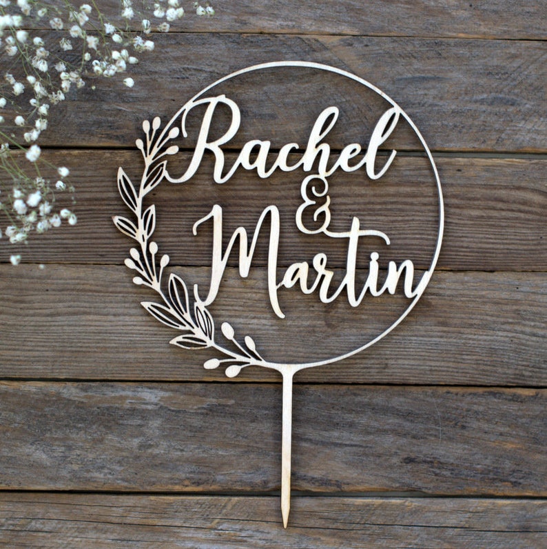May include: A wooden cake topper with the names "Rachel & Martin" in elegant script, encircled by a floral wreath. The topper is designed for weddings or anniversaries, with a stake for easy insertion into a cake.
