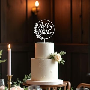 May include: A two-tiered white wedding cake with a floral arrangement and a cake topper. The cake topper is a white circular design with the names "Ashley & Heather" in script, surrounded by a floral wreath. A lit candle is in the background.