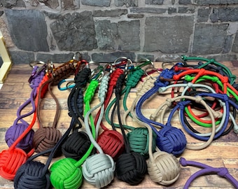 May include: A collection of colorful knotted keychains with various colored cords and metal key rings. The keychains are tied into a ball shape and are displayed on a wooden surface.