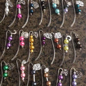 May include: A collection of 20 silver bookmark pendants with various charms and beads. The charms include a crescent moon, a star, a cross, and a variety of other designs. The beads are made of glass and come in a variety of colors, including purple, pink, green, blue, and gold.