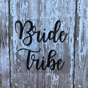 May include: Black glitter vinyl decal with the words "Bride Tribe" on a whitewashed wood background.