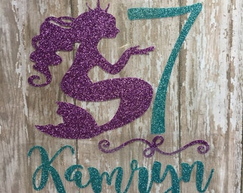 May include: Purple glitter mermaid silhouette with a teal glitter number 7 and the name Kamryn in teal glitter.