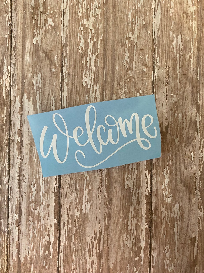 Op de afbeelding: Een lichtblauwe vinyl sticker met het woord "Welkom" in witte cursieve letters.