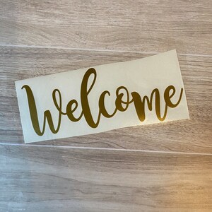 Op de afbeelding: Gouden vinyl sticker met het woord "Welkom" in een cursief schrift.